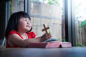 Little girl praying and holding wooden cross on bible at home, child's pure faith, focus at face. photo