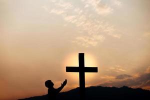 silueta de un hombre rezando frente a la cruz en la montaña al atardecer. concepto de religión. foto