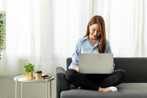 feliz mujer asiática casual y hermosa que trabaja en una computadora portátil sentada en un sofá como independiente, trabaja desde el concepto de casa. foto