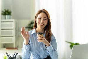 Asian woman sitting at wooden desk in office, looking at camera, waving hello. photo