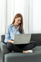 feliz mujer asiática casual y hermosa que trabaja en una computadora portátil sentada en un sofá como independiente, trabaja desde el concepto de casa. foto