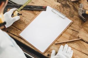 Mockup blank clipboard with white sheet and pencil on carpenter table, Furniture designer holding tape measure planning project at workbench, Man making notes in woodworking shop photo