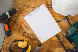 Mockup blank clipboard with white sheet and pencil on carpenter table, White safety helmets and worktools on workbench in woodworking shop photo