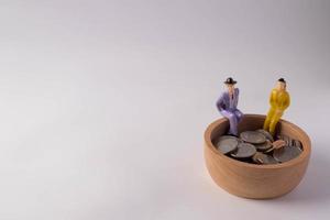 Miniature people. Sitting on a wooden bowl with coins on a white background. Money saving concept. Copy space. photo