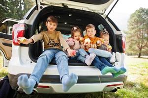 Family of four kids eat apples at vehicle interior. Children sitting in trunk. Traveling by car in the mountains, atmosphere concept. photo