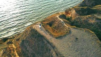 Young couple standing close, hugging on high hill of seashore in sun rays. Aerial view from drone of man and woman in love standing on cliff above sea surface in sunset light. Concept of romance video