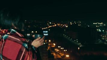 vista lateral de una joven mujer bonita mirando la ciudad nocturna, sosteniendo una taza de té, relajándose por la noche. mujer de cabello oscuro envuelta en una manta a cuadros, tomando una bebida caliente por la noche. concepto de estilo de vida video