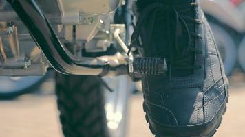 Crop view of male foot in leather shoe pushing lever of motorcycle, trying to start motorbike on road. Biker pressing on gas pedal by foot, getting bike in balance outdoor. Concept of motorcycling video