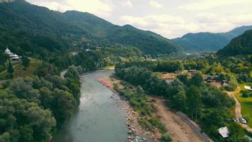 Meandering river running among dense forest and mountains. Aerial drone view of mountain river flowing between hills and small villages in Carpathians, Ukraine. Concept of landscape video