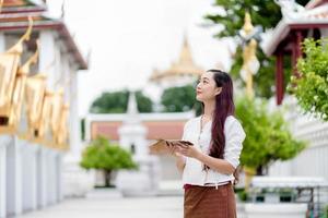 Asia woman wearing traditional dress of Thailand looking temple and hold Sanskrit ancient Tripitaka book of Lord Buddha dhamma, Sanctuary Ratchanatdaram bangkok. photo