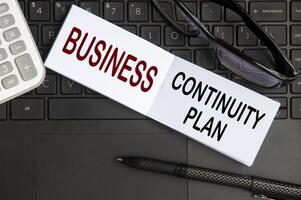 Business continuity plan text on notepad with glasses, pen and laptop background. Business concept photo