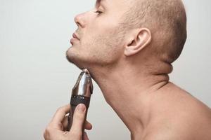 Cropped image side view of a young bearded man using electric shaver isolated over gray background. photo