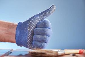 Hand wearing blue work glove with thumb up on blue background and against the background of painted wooden surface.