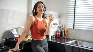 hermosa joven haciendo y bebiendo batidos de frutas en la cocina en casa mientras escucha música a través de auriculares - conceptos de estilo de vida foto