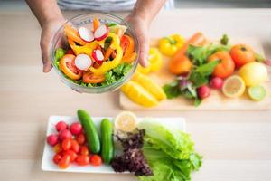 close-up top view of a vegetable salad holding in hand against a blurred background of vegetables on the table such as tomatoes, cucumbers, green oak, red oak, lemon in the kitchen. photo