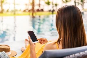 Young woman traveler relaxing and using a mobile phone by a hotel pool while traveling for summer vacation photo