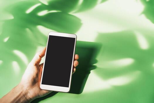 mano sosteniendo un teléfono inteligente de pantalla vacía con sombra de licencia verde tropical sobre fondo de color con espacio de copia, vista superior foto