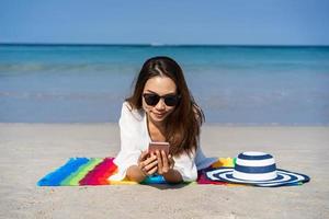 Young asian woman traveler using her smartphone and sunbath at tropical sand beach, Summer vacation concept photo