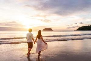 Young woman traveler holding man's hand and looking beautiful sunset on the beach, Couple on vacation in summer concept photo