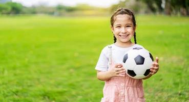 niña sonriente sosteniendo un balón de fútbol de pie en el campo de fútbol verde en el día de verano. retrato de niña atleta jugando con una pelota en el estadio. concepto de infancia activa. copie el espacio foto