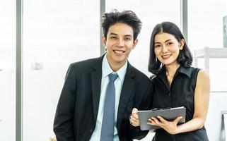 Professional asian business man in a suit and a woman in a black dress standing in the office looking at the camera with confident smiles at the success of their partnership, holding a file photo