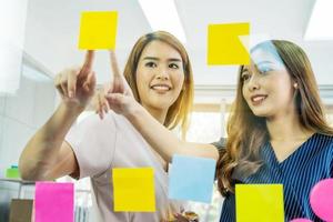 Two young business woman pointing sticky note papers on the glass thinking new idea with colleague photo