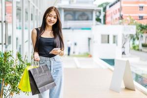 Smiling asian girl is holding shopping bags, using a smartphone while doing shopping in the mall, shopping concept, portrait, smiling. photo