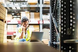 Asian female worker in safety vest sitting and working with computer laptop in storage warehouse. She having a lot hard work and many trouble per day photo