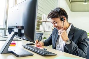 el centro de llamadas masculino asiático con auriculares se sienta en la mesa consulta al cliente en línea y toma notas en la hoja de texto mientras habla por teléfono foto