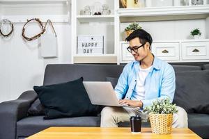 Smart Asian businessman in casual clothing sitting on a sofa in the house is enjoying the work by using the computer laptop to make money from online business, work from home photo