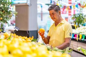 Old senior asian man looking at tangerine while enjoying grocery shopping at in the supermarket photo