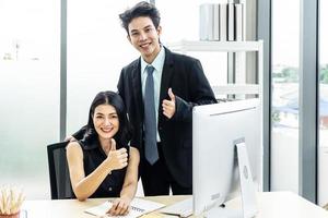 hombres de negocios alegres, dos hombres de negocios y una mujer sentados y parados en la mesa, dos negocios mirando la cámara y mostrando el pulgar hacia arriba foto