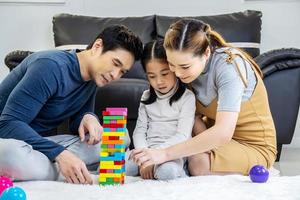 Happy asian family father and mother with little asian girl smiling playing with building tower from wooden blocks, taking tiles in turn from underneath until it falls, in moments good time at home photo