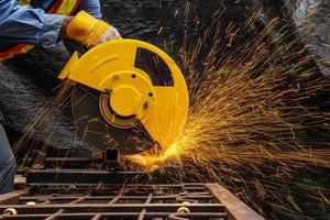 ingeniero técnico industrial en tela protectora azul con máscara de seguridad, hombre que trabaja con una herramienta de amoladora eléctrica en una estructura de acero con chispas volando en la fábrica, enfoque selectivo foto