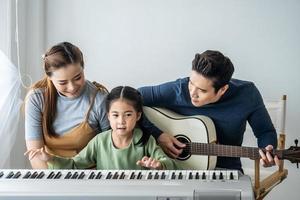 feliz pequeña hija asiática tocando el piano con madre y padre tocando la guitarra en casa, madre enseñando a su hija a tocar el piano, tocan y cantan canciones. Ellos se están divirtiendo. foto