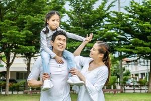 Portrait of asian happy family of three having fun together in park,  little daughter sitting on father back and mother smile walking and having fun moments good time in summer park at home photo