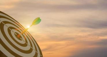 Silhouette of green dart arrow hitting bullseye target center dartboard on sunset background. Business targeting and focus concept, strategy, achievement, and planning concept photo