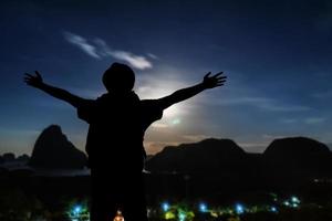 siluetas de turistas, fotógrafos de pie con una mano feliz en la cima de una montaña alta. mira felizmente las estrellas en el cielo y la vía láctea en la noche. Larga exposición con grano. foto