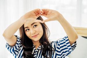 Beautiful asian young woman sitting on bed and making heart shape with fingers. Portrait of smiling lady posing in her bedroom, Concept happiness and love. photo