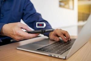Businessman holding a phone with a low battery charge screen While working on the laptop , I need to find a quick charger photo