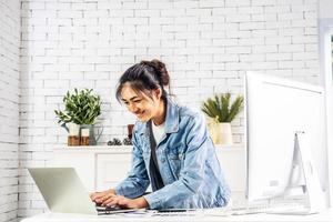 joven mujer asiática feliz sonriente relajándose conectada en Internet con computadora portátil trabajando y reunión de videoconferencia en casa. joven escribiendo creativo en el teclado, concepto de trabajo desde casa foto