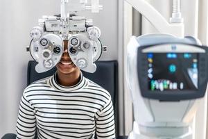 Una joven sonriente afroamericana con cabello afro haciendo una prueba ocular en un foróptero óptico, comprobando su ojo con una máquina de optometría foto