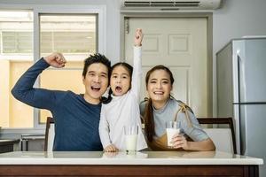 Happy family having fun in the kitchen. Asian Father, mother and little daughter spending time together and having breakfast drinking and hold glasses of milk at table photo