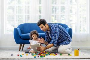 concepto moderno de comunicación remota. retrato de un padre caucásico positivo haciendo una videollamada virtual con su madre usando una tableta, mientras la pequeña hija jugaba con coloridos bloques de madera foto