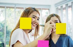 las mujeres de negocios creativas asiáticas están escribiendo una nota en la hoja de texto después de pensar en una nueva idea y planificar un proyecto con un colega, un grupo de empleados con una lluvia de ideas sobre una pared de vidrio y pegajosa foto