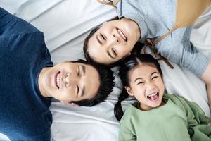 vista superior de la hermosa joven madre asiática feliz, el padre y su hija mirando a la cámara y sonriendo mientras están acostados en la cama cabeza a cabeza en el dormitorio foto