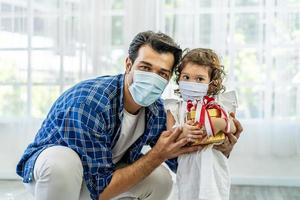 Happy father and daughter in medical mask holding gift box. Christmas online holiday celebration, Xmas, new year in lockdown coronavirus covid-19 quarantine. New normal, social distance, stay home photo