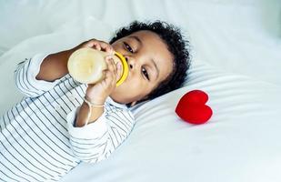 African american cute baby boy drinking milk from bottle and sleeping on bed with a red heart at home, Children need love and caring