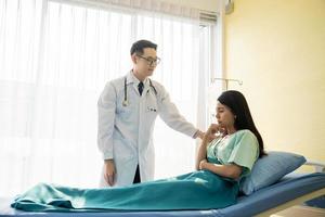 médico masculino asiático usando estetoscopio y consolando a una paciente infeliz sentada en una cama de hospital foto