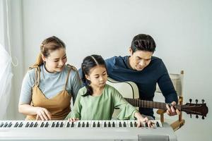 feliz pequeña hija asiática tocando el piano con madre y padre tocando la guitarra en casa, madre enseñando a su hija a tocar el piano, tocan y cantan canciones. Ellos se están divirtiendo. foto